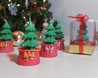 Könnte beinhalten: Vier grüne Weihnachtsbaumkerzen in roten Töpfen mit goldenen Schleifen. Die Kerzen befinden sich in einer durchsichtigen Schachtel mit rotem Band und goldenen Verzierungen.