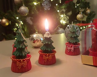 Könnte beinhalten: Drei grüne Weihnachtsbaumkerzen mit roten und goldenen Sockeln. Die Kerzen sind angezündet und die Flammen sind sichtbar. Die Kerzen stehen auf einer weißen Oberfläche.