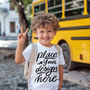May include: A young boy wearing a white t-shirt with the text "place your design here" in black. He is standing in front of a yellow school bus and giving a peace sign.