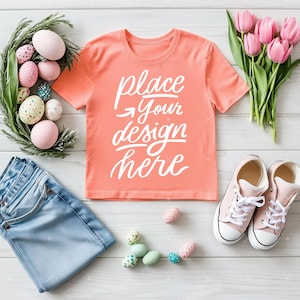 May include: A coral-colored t-shirt with the text "place your design here" in white script. The shirt is laid out with light blue jeans, pink sneakers, and Easter eggs. A bouquet of pink tulips and a green wreath with eggs complete the spring theme.