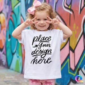 May include: A white t-shirt with the text "place your design here" in black script. The child is wearing pink trousers with a leopard print and a pink bow. The background is a colourful graffiti wall.