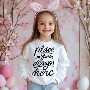 Op de afbeelding: Een wit sweatshirt met de tekst "place your design here" in zwarte letters. Een jong meisje draagt het sweatshirt en heeft een roze hoofdband op.