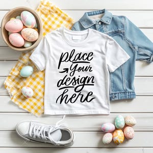 Może przedstawiać: Biała koszulka z napisem "place your design here" czarnymi literami. Koszulka leży na białym drewnianym tle z żółto-białą kratą, dżinsową kurtką, parą białych butów sportowych i miską kolorowych jajek wielkanocnych.