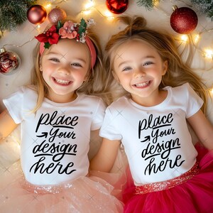 May include: Two young girls wearing white t-shirts with the text "place your design here". They are wearing pink and red tutus and flower headbands. Christmas ornaments and lights are in the background.