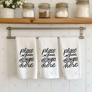 May include: Three white kitchen towels hanging on a silver rack. Each towel has the text "place your design here" written in black.