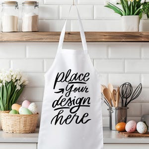 Op de afbeelding: Witte schort met zwarte tekst die "place your design here" zegt. De schort hangt in een keukenomgeving.