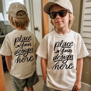 May include: Two children wearing cream-coloured t-shirts with the text "place your design here" in black script. One child wears sunglasses and a tan cap. The other child wears a tan cap. Both are wearing shorts.