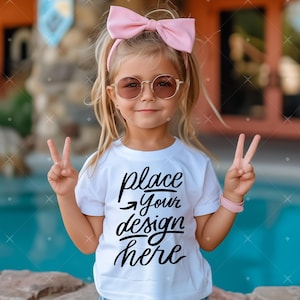 Op de afbeelding: Een jong meisje draagt een wit T-shirt met de tekst "place your design here" in zwart. Ze draagt een roze zonnebril en een roze strik in haar haar. Ze maakt met beide handen het vredesteken.