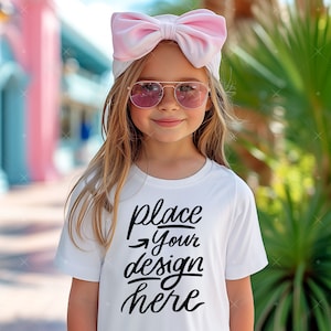 Könnte beinhalten: Ein junges Mädchen trägt ein weißes T-Shirt mit dem Text "place your design here" in schwarzen Buchstaben. Sie trägt ein rosa Stirnband und eine Sonnenbrille.