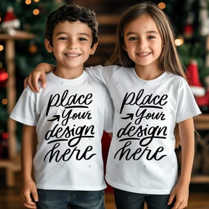 Op de afbeelding: Twee kinderen dragen witte t-shirts met de tekst "place your design here" in zwart schrift. De kinderen lachen en hebben hun armen om elkaar heen. De achtergrond is wazig met kerstversieringen.