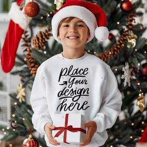 Op de afbeelding: Een kind draagt een kerstmuts en een witte trui met de tekst "place your design here". Het kind houdt een witte geschenkdoos met een rode strik vast voor een versierde kerstboom.