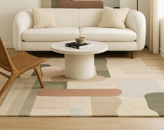 May include: A modern living room featuring a cream-colored sofa, a round coffee table, and a patterned rug. A large abstract wall hanging in neutral tones complements the decor. A woven chair and a black floor lamp add to the room's design.