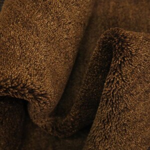 May include: Close-up of a folded, brown, plush fabric. The material has a soft, textured surface with a dense, fuzzy pile. The fabric is a warm, earthy tone, with visible folds and shadows.