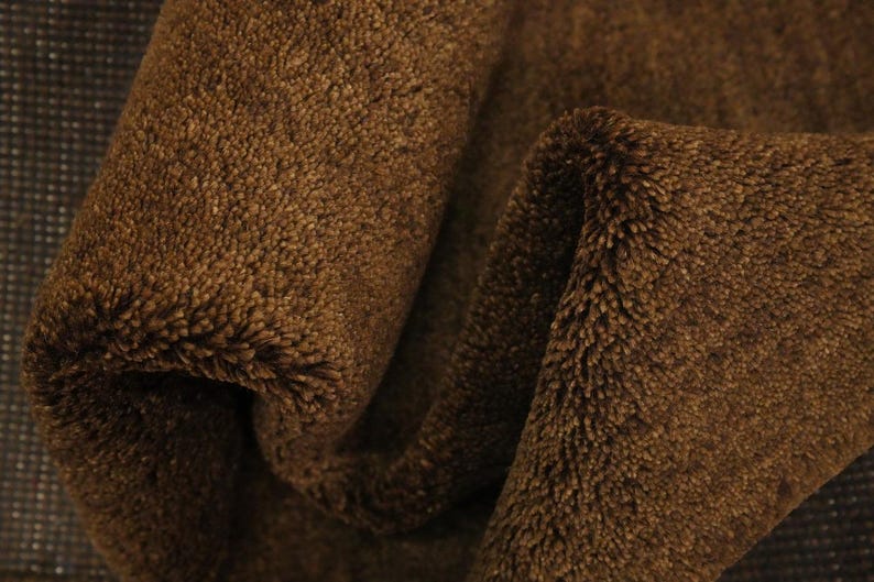 May include: Close-up of a folded, brown, plush fabric. The material has a soft, textured surface with a dense, fuzzy pile. The fabric is a warm, earthy tone, with visible folds and shadows.
