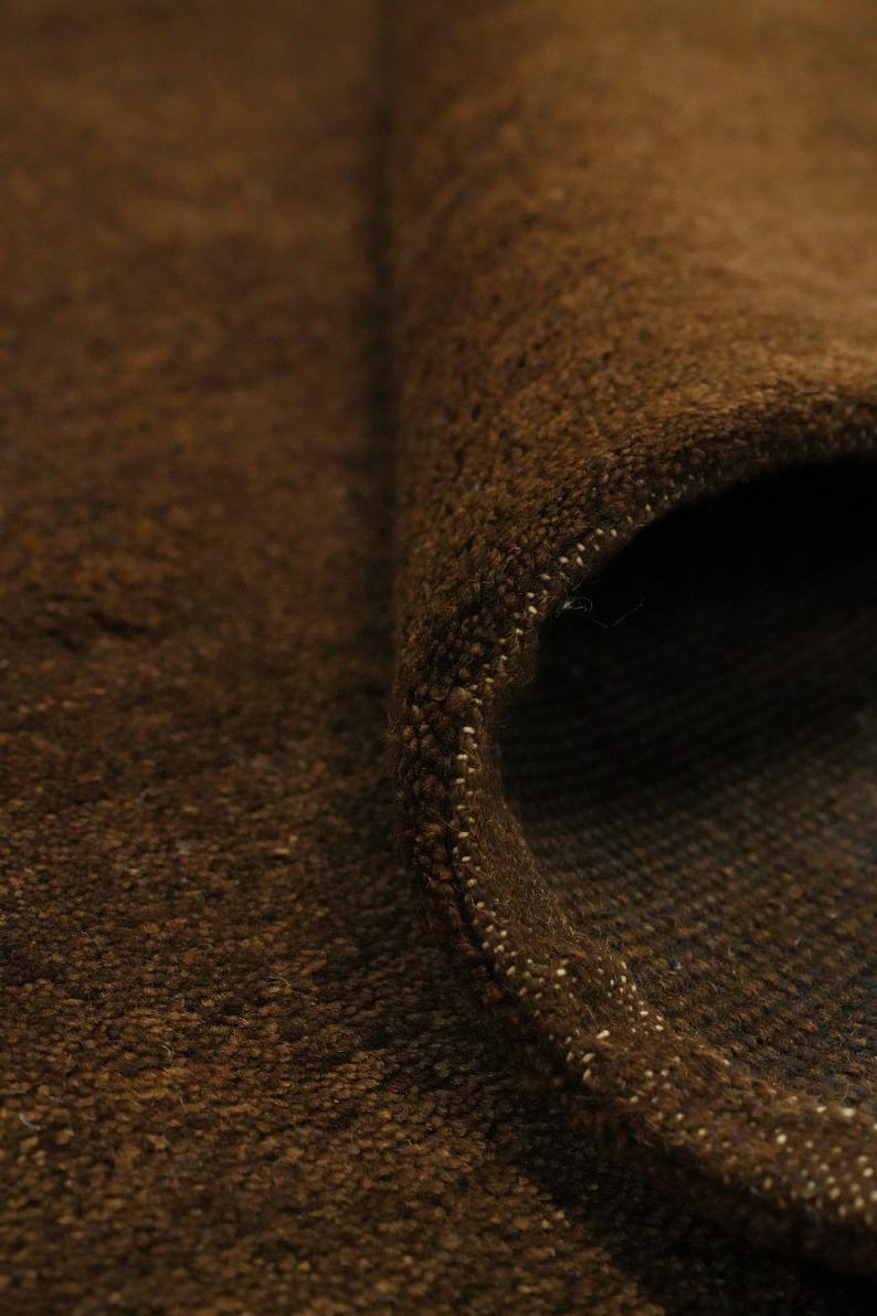 May include: Close-up of a rolled-up brown rug. The rug has a textured surface with visible fibers and a rolled edge. The color is a rich, warm brown, with a hint of lighter flecks along the edge. The rug appears to be made of a thick, durable material.