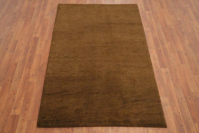 May include: A rectangular brown rug with a textured surface. The rug is placed on a wooden floor with a warm, reddish-brown tone. The rug's color is a solid, earthy brown.