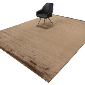 Puede incluir: Una alfombra grande, rectangular, marrón con un borde marrón más oscuro. Una silla negra con patas plateadas está sobre la alfombra. La alfombra tiene un aspecto texturizado y es adecuada para una sala de estar o un dormitorio.