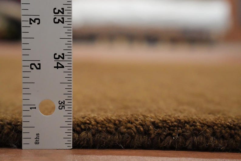 May include: Close-up of a brown rug with a ruler showing the pile height. The rug has a dark brown edge and a lighter brown surface. The ruler indicates the rug is approximately 33 inches long.