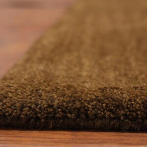 May include: Close-up of a brown rug with a textured surface. The rug's edge is visible, showing the thickness and density of the fibers. The rug is placed on a wooden floor.
