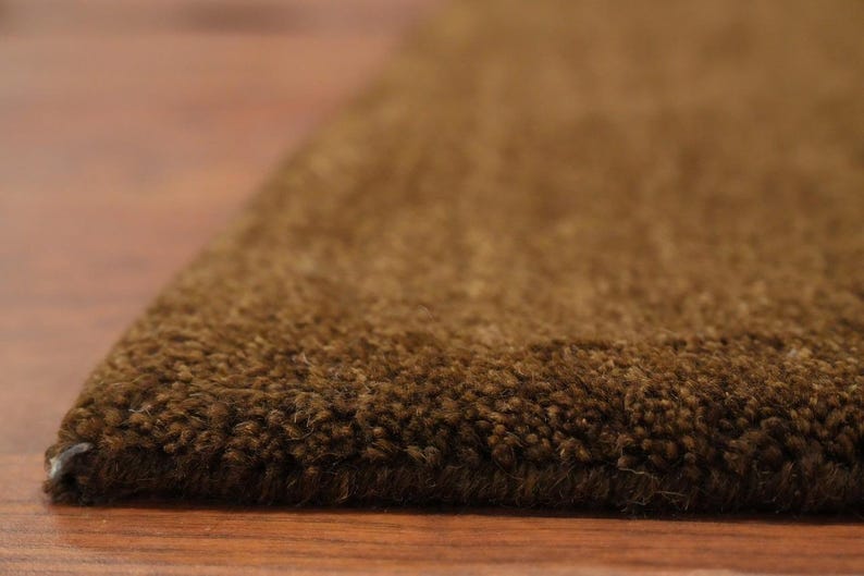May include: Close-up of a brown rug with a textured surface. The rug's edge is visible, showing the thickness and density of the fibers. The rug is placed on a wooden floor.