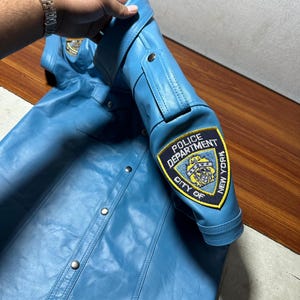 以下が含まれることがあります： ボタン留めのフロントと襟が付いた、ライトブルーの革のシャツ。袖には「POLICE DEPARTMENT CITY OF NEW YORK」のパッチが付いています。シャツはシルバーの金属製ハンガーで吊るされています。