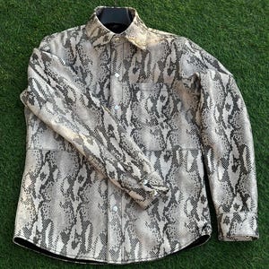 May include: A snakeskin-patterned shirt in grey, black, and cream tones. The long-sleeved shirt features a collar, a chest pocket, and silver buttons. It is displayed on a black hanger against a green backdrop.