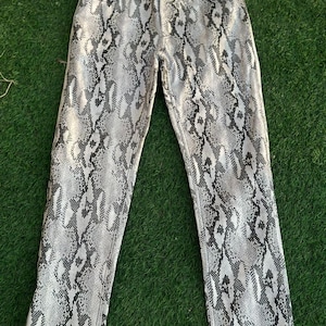 May include: Straight-leg jeans featuring a snakeskin print in grey and black. The trousers are displayed flat on a green surface, highlighting the full length and pattern of the fabric.