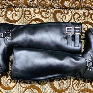 May include: A pair of black leather knee-high boots with silver-tone metal hardware. The boots have buckle fastenings and a thick, lug sole. The boots are displayed on a patterned surface.