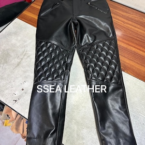 Op de afbeelding: Zwarte leren broek met gewatteerde kniepanelen en zakken met ritssluiting. De broek heeft een knoopsluiting en riemlussen. De tekst "SSEA LEATHER" is op de broek gedrukt.