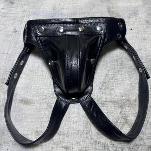 May include: Black leather jockstrap with silver-tone rivet details. The jockstrap features a pouch and adjustable straps. The design is made of smooth, shiny leather.