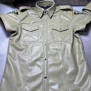 May include: A tan-coloured, short-sleeved shirt with a collar, two chest pockets, and silver-coloured buttons. The shirt has shoulder patches with text. The stitching is visible, and the shirt appears to be made of a leather-like material.