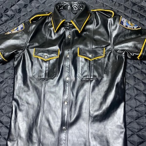 May include: Black leather short-sleeve shirt with yellow trim on the collar, pockets, and sleeves. Features two chest pockets and silver buttons down the front. Includes a patch on each sleeve that reads "POLICE DEPT CITY OF NEW YORK".