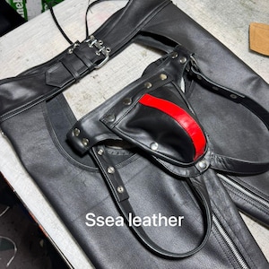 Puede incluir: Chaps de cuero negro con una raya roja y detalles plateados. Los chaps tienen cierre de cremallera y correas ajustables. El texto "Ssea leather" es visible.