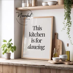 Könnte beinhalten: Ein gerahmter Druck mit dem Text "This kitchen is for dancing" in einer Küchenumgebung. Der Druck ist gegen eine Holzoberfläche gelehnt, mit einem Schneidebrett, Schalen und einer kleinen Topfpflanze in der Nähe. Die Küche hat eine helle und luftige Atmosphäre.