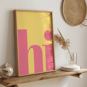 Peut inclure: Impression d'art encadrée avec le mot "hi" en rose sur fond jaune. L'œuvre a un design minimaliste et est exposée sur une étagère en bois. Le cadre est de couleur marron clair.