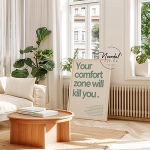 May include: A light wood round coffee table with a stack of books on top. A framed poster with the text "Your comfort zone will kill you" is next to the table. A large green plant and a white sofa are also in the room.