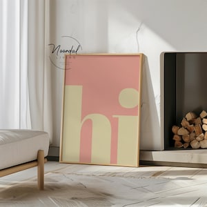 May include: A framed art print with the word "hi" in a retro font. The background is a soft pink, with the letters in a creamy beige. The frame is a light wood color. The print is leaning against a wall near a fireplace.