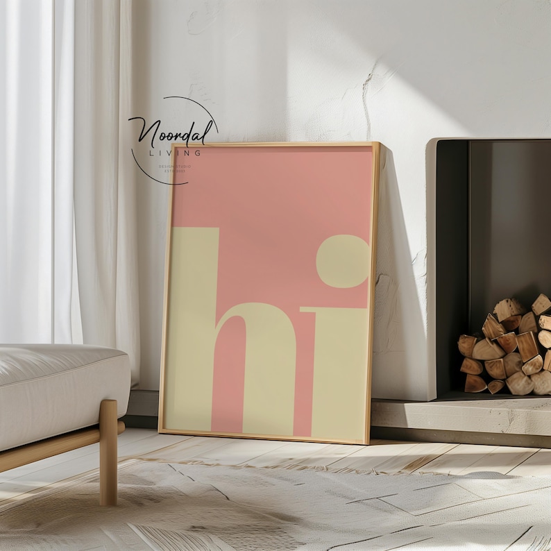 May include: A framed art print with the word "hi" in a retro font. The background is a soft pink, with the letters in a creamy beige. The frame is a light wood color. The print is leaning against a wall near a fireplace.