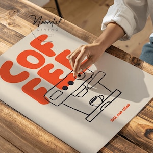 May include: A cream-colored poster with the word "COFFEE" in large orange letters. Below the word is a black and white illustration of a coffee machine. The words "RISE AND GRIND" are printed in orange. The poster is on a wooden table.