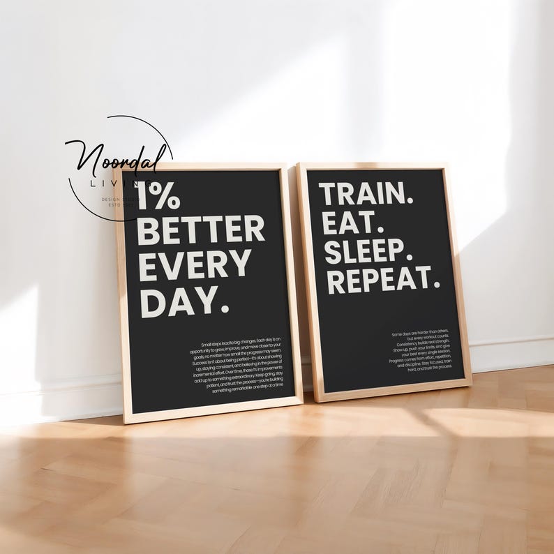 Two black framed typography prints leaning against a white wall on a wooden floor, with one reading 1% better every day and the other train eat sleep repeat.