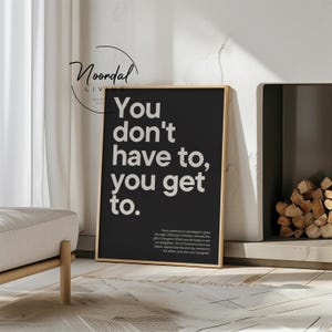 A black framed poster with white text reading "You Don't Have To, You Get To" and a smaller paragraph of text below, leaning against a white wall next to a fireplace.