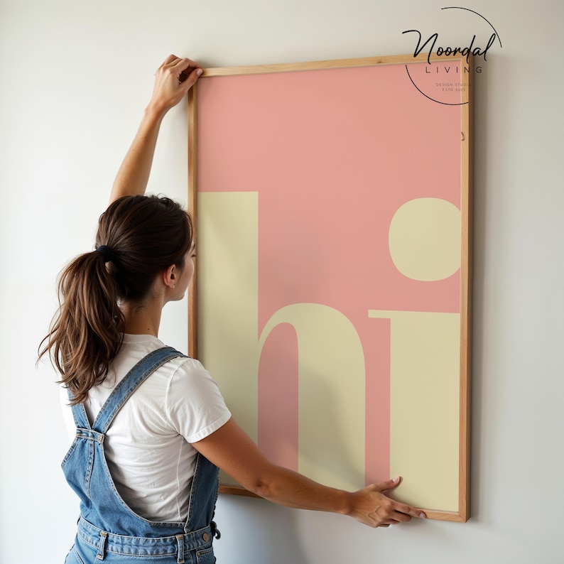 May include: A framed art print with the word "hi" in a large, stylized font. The background is a soft pink, with the letters in a creamy yellow. The frame is a light wood color. The print is being held up by a person.