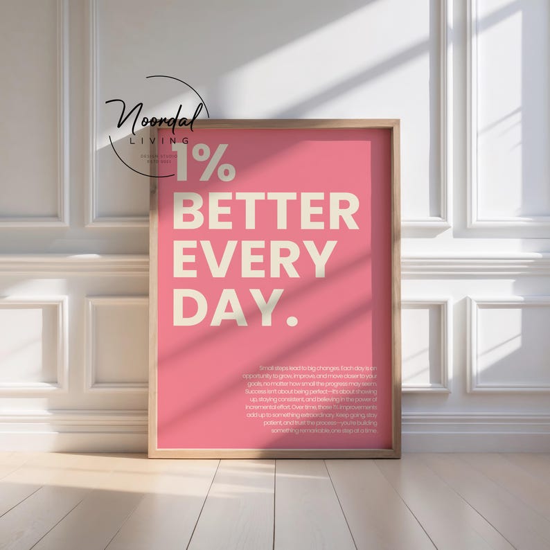 A framed pink motivational poster leaning against a white paneled wall on a wooden floor, featuring cream-colored text reading 1% better every day with a small paragraph underneath.