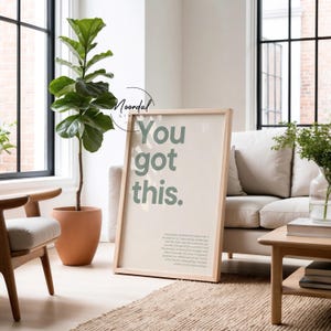 Könnte beinhalten: Ein gerahmter Druck mit dem Text "You got this" in hellgrüner Farbe. Der Druck befindet sich in einem hellen Holzrahmen und lehnt an einem beigen Sofa. Links befindet sich eine große grüne Pflanze in einem Terrakottatopf.