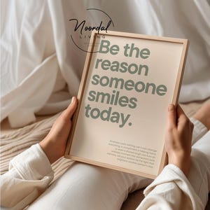 Könnte beinhalten: Ein rechteckiger Holzrahmen mit einem beigen Druck und dem salbeigrünen Text Be the reason someone smiles today. Unter dem Haupttext steht in kleinerer salbeigrüner Schrift: Kindness costs nothing, yet it can change everything.