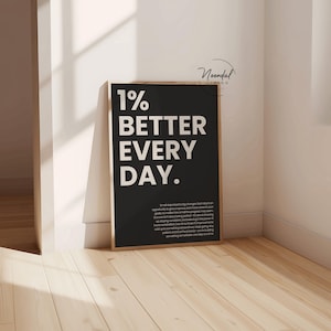 A single black framed print reading 1% better every day standing on a wooden floor in a corner with white walls.