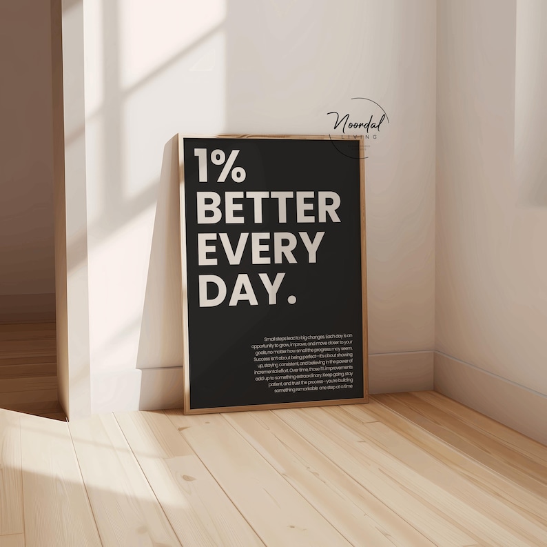 A single black framed print reading 1% better every day standing on a wooden floor in a corner with white walls.