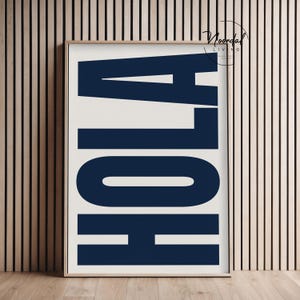Op de afbeelding: Een ingelijste kunstprint met het woord "HOLA" in grote, marineblauwe blokletters op een gebroken witte achtergrond. Het kunstwerk zit in een houten lijst en wordt getoond tegen een houten lattenwand.