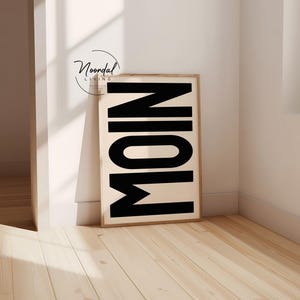 May include: A framed print featuring the word "MOIN" in large, black letters on a cream-colored backdrop. The wooden frame adds a touch of warmth. The artwork is propped against a white wall on a light wood floor.