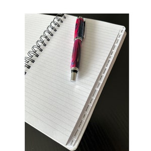 Może przedstawiać: Szyty spiralnie notes z czarno-srebrnym długopisem na górze. Notes posiada zakladki z następującymi etykietami: MISC, PHARM, LAB/RAD, URO, RHEUM, PULM, OTOJOPH, ORTHO, NEURO, NEPRO, MENT, LA, NLTH, NEME, MOTION.